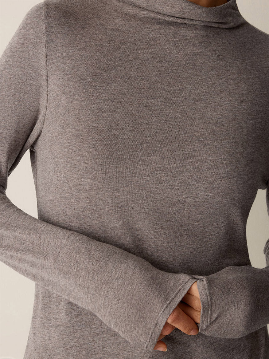 Wearing the Eileen Fisher Mélange Sheer Cotton Blend Knit Funnel Neck Top in Cobblestone, this person shows off the long sleeves with thumbhole cuffs and a hand partially tucked inside—the perfect lightweight layer for any season.