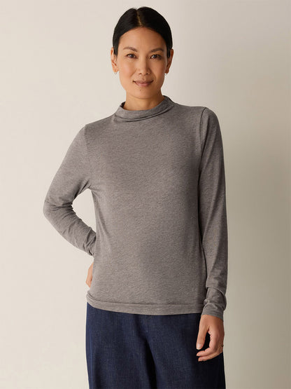 A person stands against a plain background, wearing the Eileen Fisher Mélange Sheer Cotton Blend Knit Funnel Neck Top in Cobblestone with dark pants, one hand on their hip and a neutral expression.