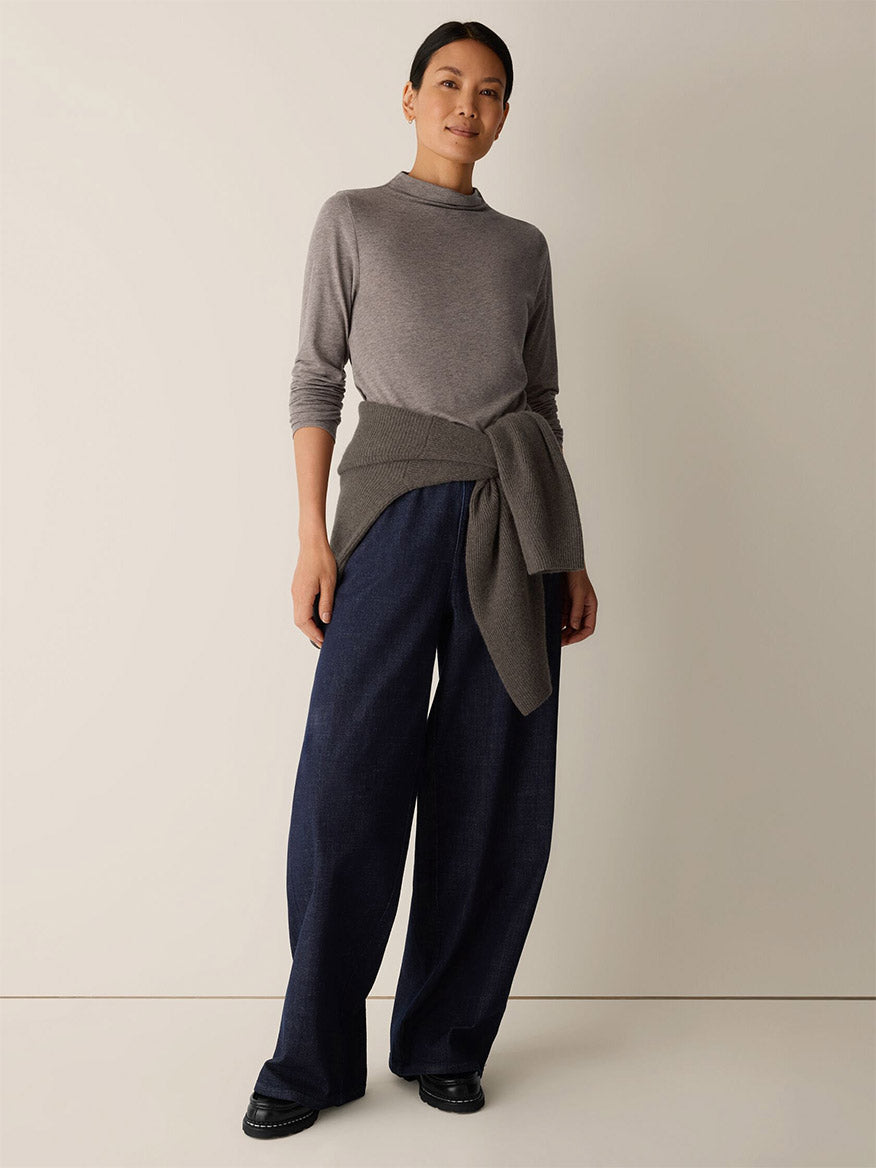 A woman stands against a plain background wearing the Eileen Fisher Mélange Sheer Cotton Blend Knit Funnel Neck Top in Cobblestone, with another grey sweater tied at her waist, wide-leg dark blue pants, and black shoes.