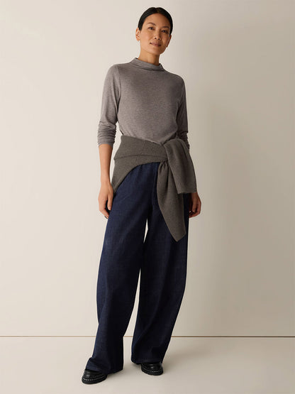 A woman stands against a plain background wearing the Eileen Fisher Mélange Sheer Cotton Blend Knit Funnel Neck Top in Cobblestone, with another grey sweater tied at her waist, wide-leg dark blue pants, and black shoes.