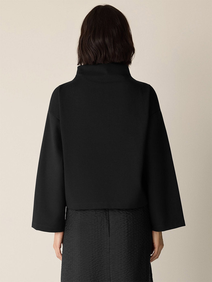 A person with shoulder-length dark hair is seen from the back, wearing an Eileen Fisher Lightweight Ponte Funnel Neck Top in black paired with a black textured skirt.
