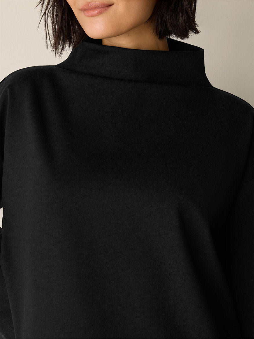 A woman is shown from the shoulders up wearing the Eileen Fisher Lightweight Ponte Funnel Neck Top in Black, featuring a high, structured neckline and a boxy fit against a neutral background.