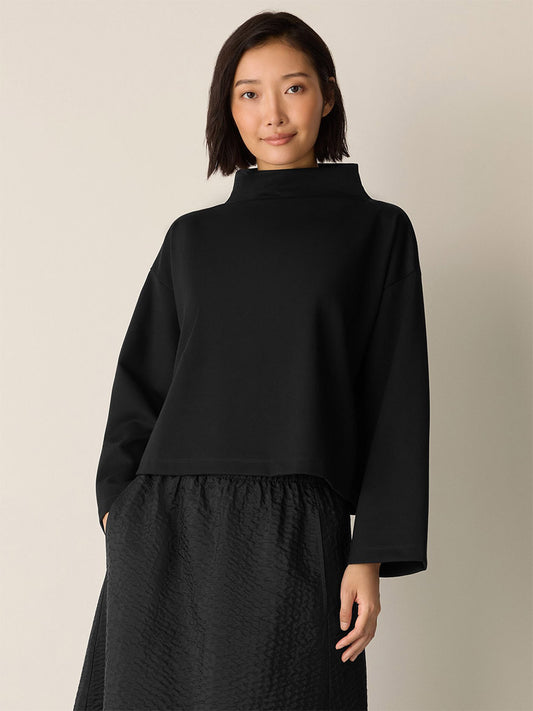 A woman with short dark hair wears the Eileen Fisher Lightweight Ponte Funnel Neck Top in Black and a textured black skirt with a boxy fit, standing against a plain light background.