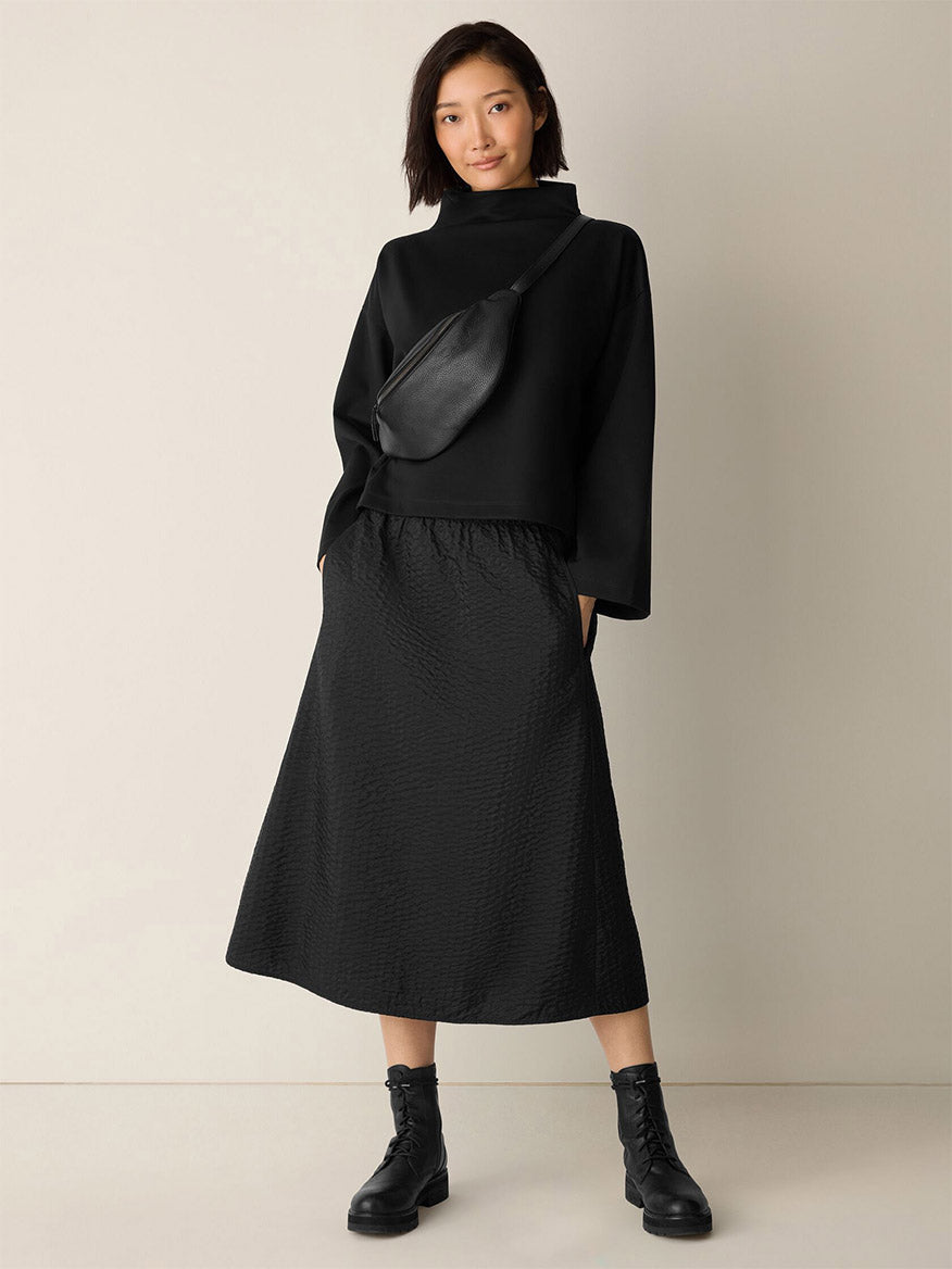 A woman stands against a plain wall wearing the Eileen Fisher Lightweight Ponte Funnel Neck Top in Black, paired with a black midi skirt, black boots, and a black crossbody bag.