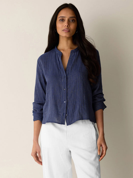 A woman with long dark hair stands against a neutral background, wearing the Eileen Fisher Striped Organic Cotton Gauze Band Collar Shirt in Deep Dive and white pants.