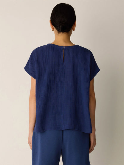 A person with dark hair tied back is shown from behind, wearing the Eileen Fisher Organic Cotton Lofty Gauze Round Neck Box-Top in Deep Dive with matching blue pants against a plain light background.