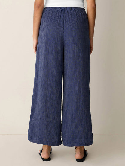 Person stands with back to camera, wearing Eileen Fisher Striped Organic Cotton Gauze Wide-Leg Pant in Deep Dive and black flat sandals, against a light-colored wall.