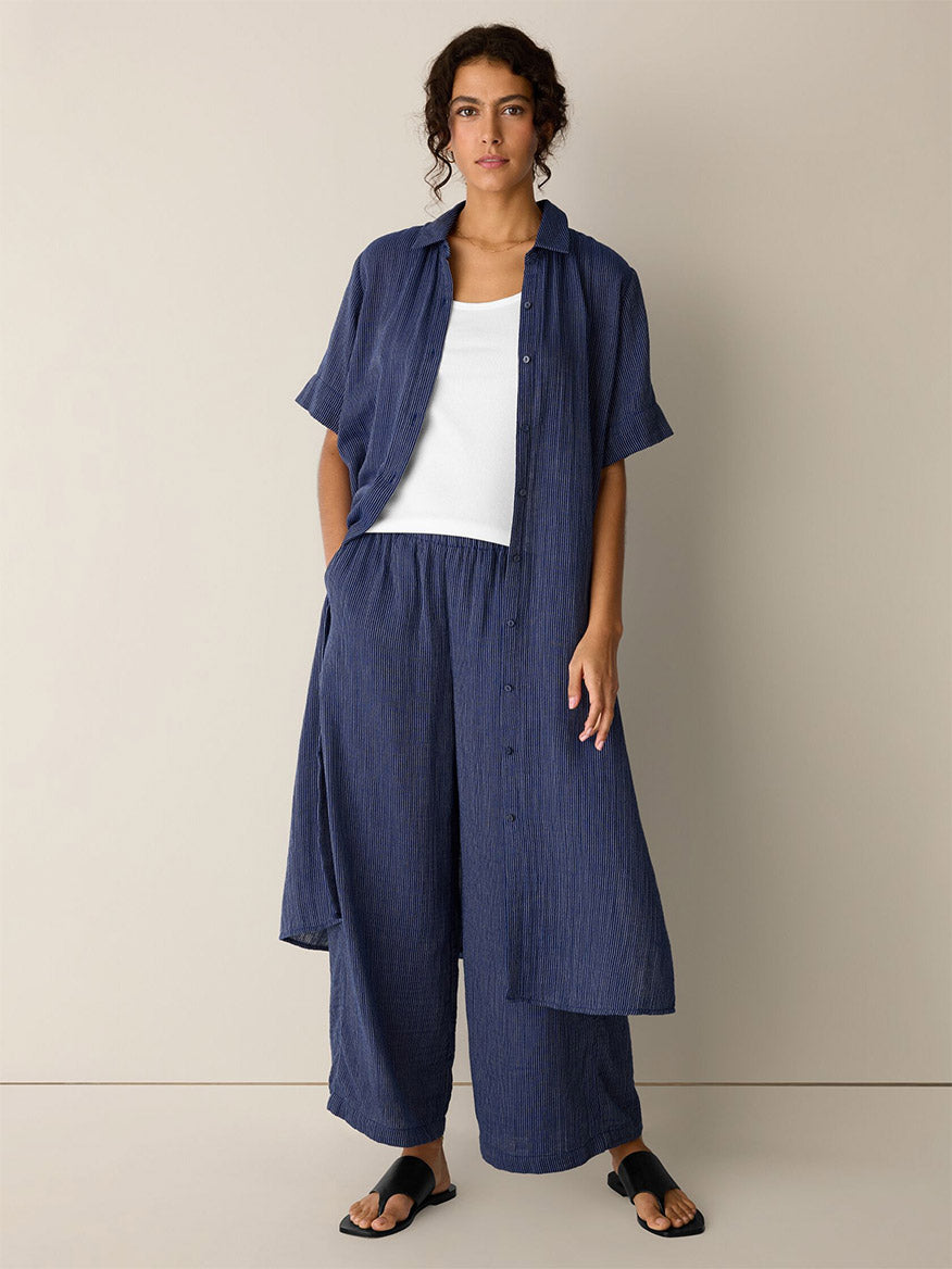 A woman wears the Eileen Fisher Striped Organic Cotton Gauze Wide-Leg Pant in Deep Dive, paired with a loose-fitting navy shirt, white tank top, and black sandals, standing against a plain beige wall.