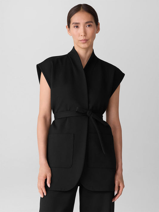 A person with long dark hair wears the Eileen Fisher Washable Flex Ponte High Collar Vest in Black, featuring short sleeves, a self-tie belt, and large front pockets, paired with matching black pants against a plain light background.