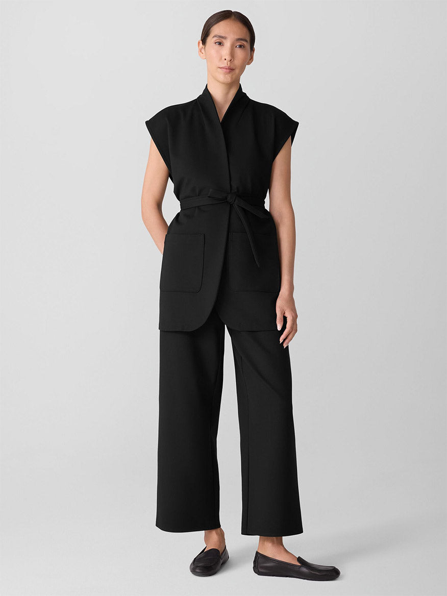 A woman stands against a plain background wearing the Eileen Fisher Washable Flex Ponte High Collar Vest in Black, styled with wide-leg black stretch ponte pants and black slip-on shoes.