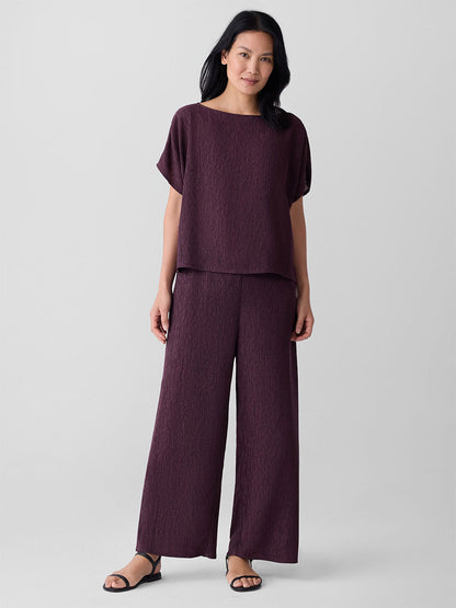 A woman stands against a plain background wearing black sandals and the Eileen Fisher Woven Plissé Wide-Leg Pant in Blackberry, paired with a loose-fitting, textured dark purple top.