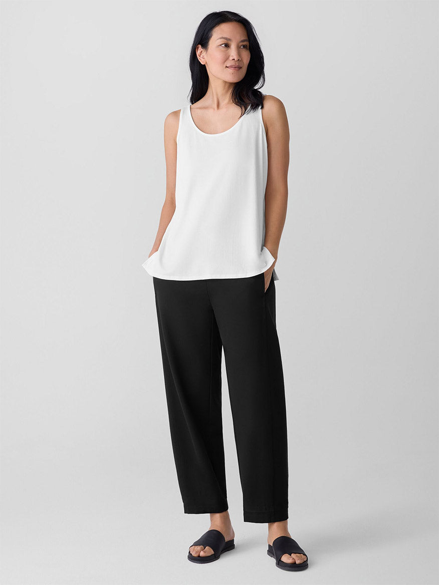 A woman stands with hands in pockets, wearing the Eileen Fisher Fine Jersey Scoop Neck Tank in White, black wide-leg pants, and black sandals against a plain light gray background.