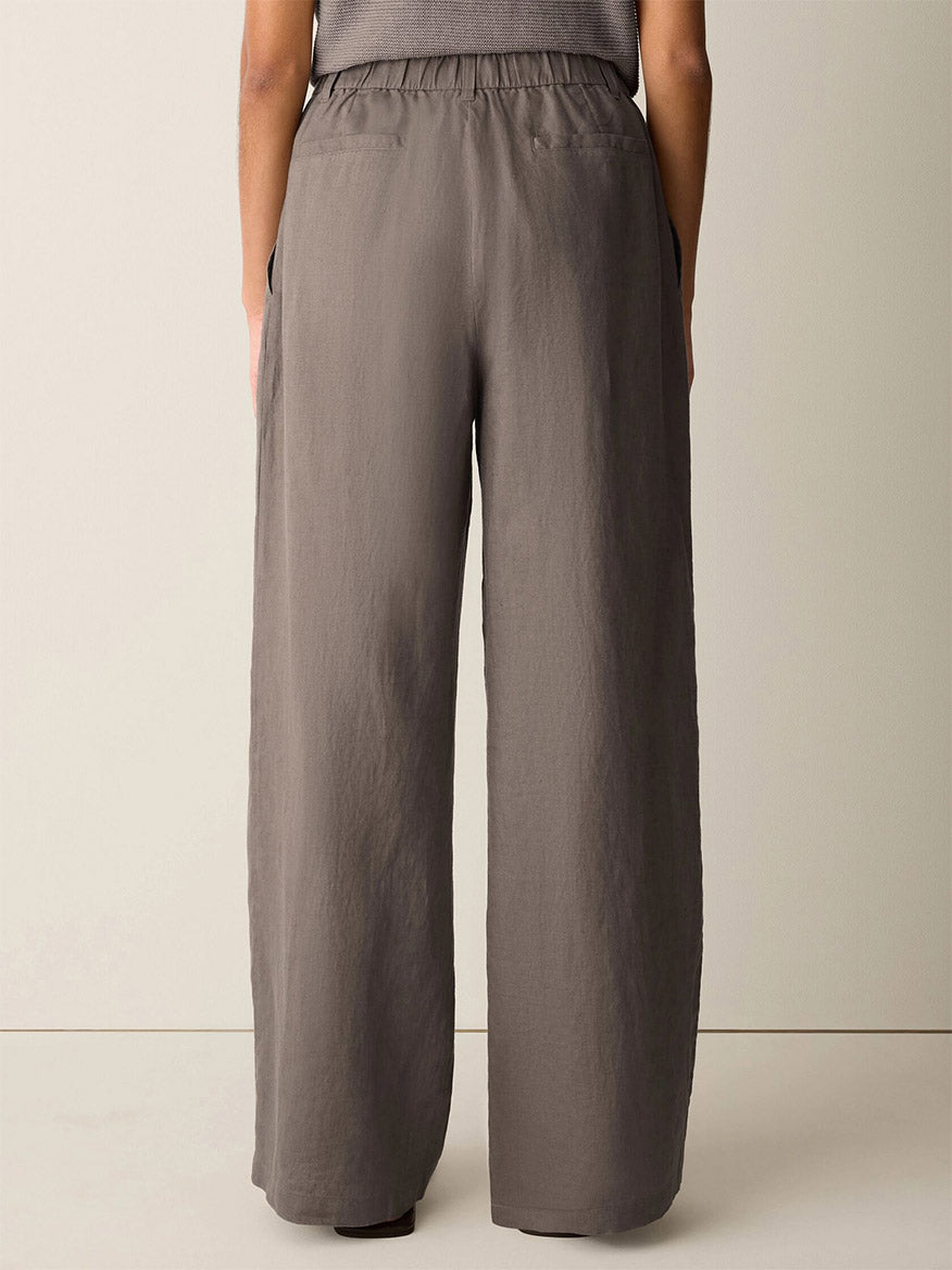A person stands facing away, wearing the Eileen Fisher Organic Linen Full-Length Trouser in Walnut—high-waisted, wide-leg pants with an elastic waistband and side pockets.