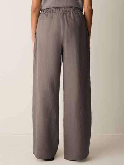 A person stands facing away, wearing the Eileen Fisher Organic Linen Full-Length Trouser in Walnut—high-waisted, wide-leg pants with an elastic waistband and side pockets.