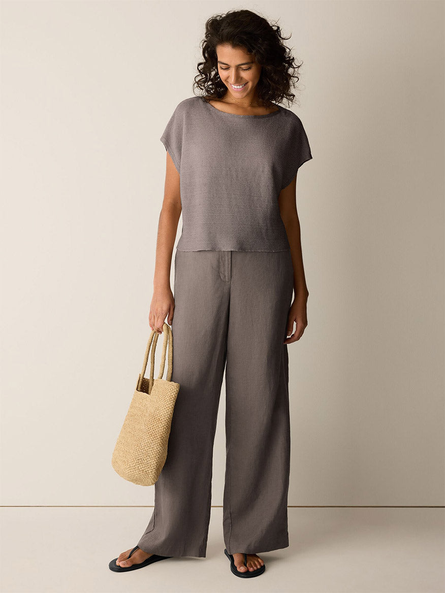 Woman standing against a plain background, wearing a loose grey organic linen top, black sandals, holding a woven straw bag, and Eileen Fisher Organic Linen Full-Length Trouser in Walnut.