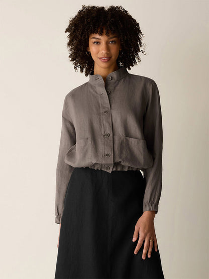 A woman with curly hair wears the Eileen Fisher Organic Linen Flight Jacket in Walnut, featuring a band collar and large front pockets, paired with a high-waisted black skirt against a plain background.