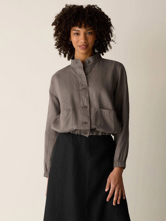 A woman with curly hair wears the Eileen Fisher Organic Linen Flight Jacket in Walnut, featuring a band collar and large front pockets, paired with a high-waisted black skirt against a plain background.