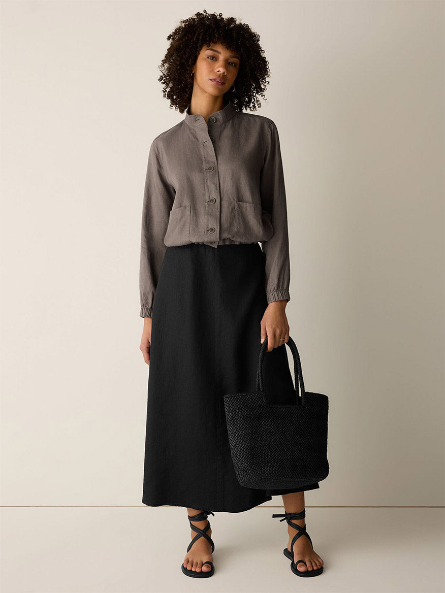 A person stands against a plain background, wearing the Eileen Fisher Organic Linen Flight Jacket in Walnut, a long black skirt, black strappy sandals, and holding a black woven tote bag.