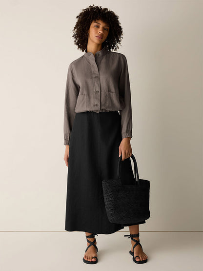 A person stands against a plain background, wearing the Eileen Fisher Organic Linen Flight Jacket in Walnut, a long black skirt, black strappy sandals, and holding a black woven tote bag.