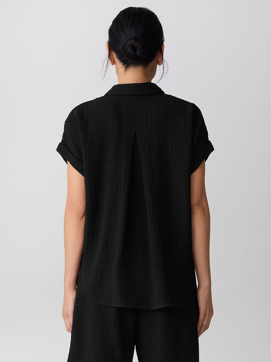 Person with dark hair in a low bun, seen from the back against a plain light background, wearing a short-sleeve black Eileen Fisher Organic Cotton Lofty Gauze Notch Collar Top paired with matching black shorts.