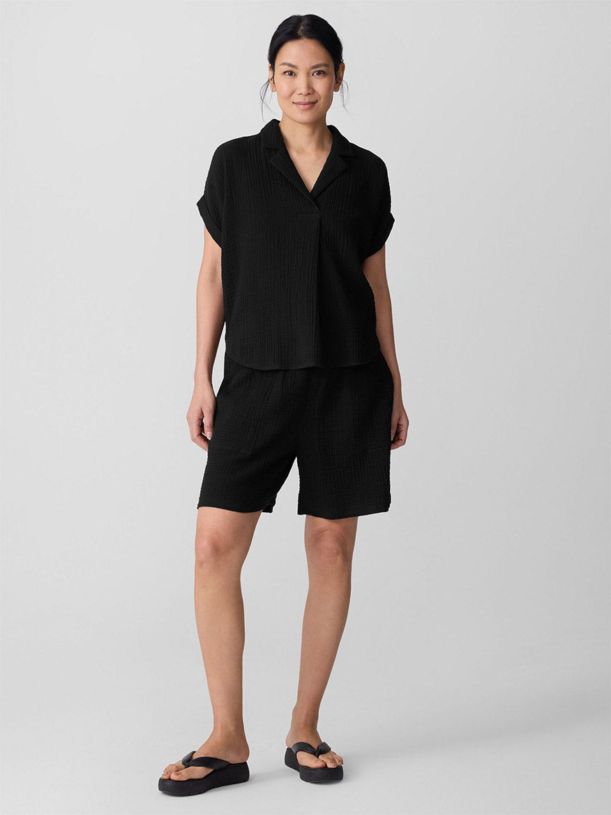 A woman stands and smiles, wearing the Eileen Fisher Organic Cotton Lofty Gauze Notch Collar Top in black with matching black shorts and sandals, set against a plain light gray background.