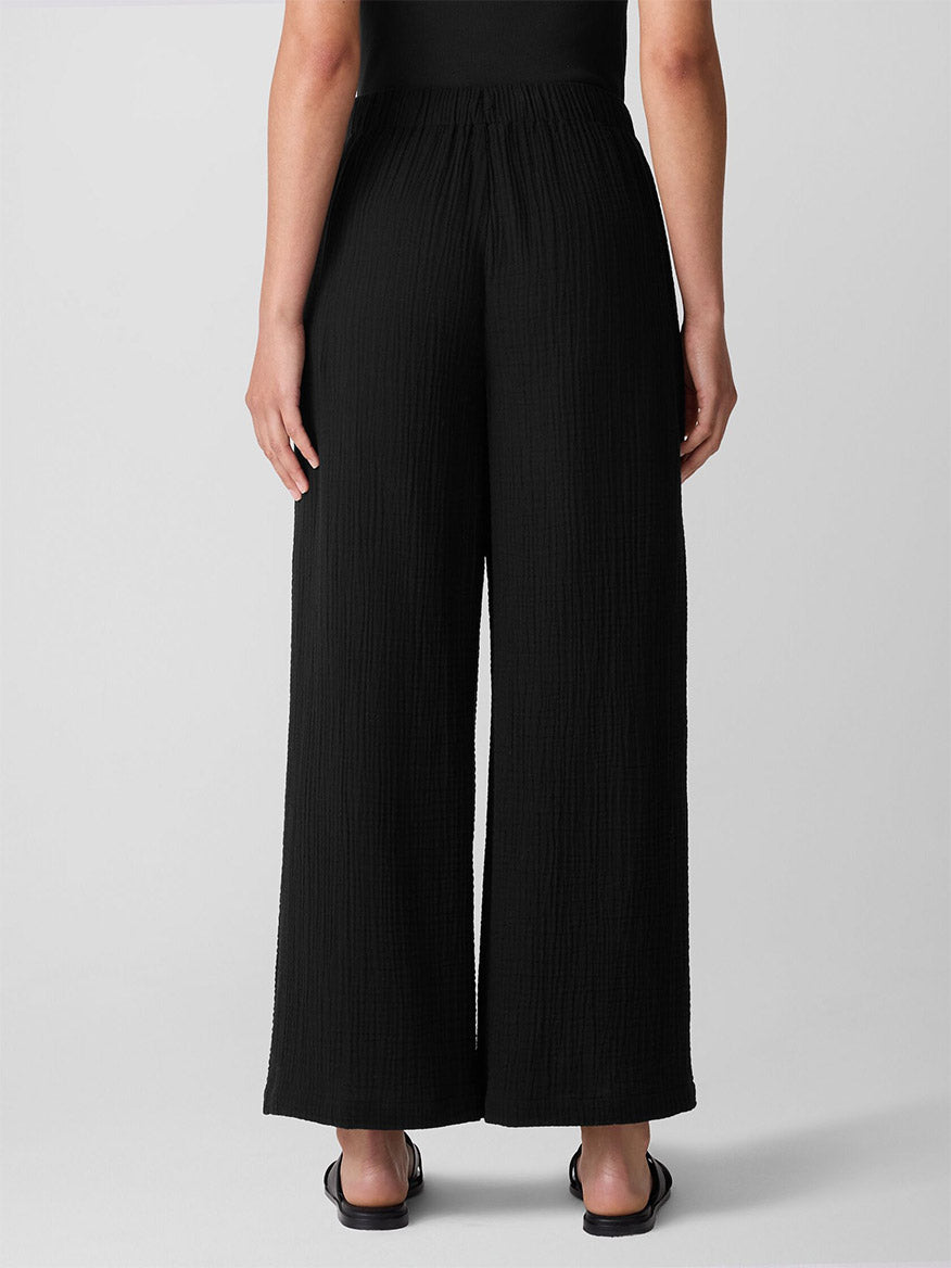 A person faces away, wearing the Eileen Fisher Organic Cotton Lofty Gauze Wide-Leg Pant in black with a matching top and sandals against a plain light background.