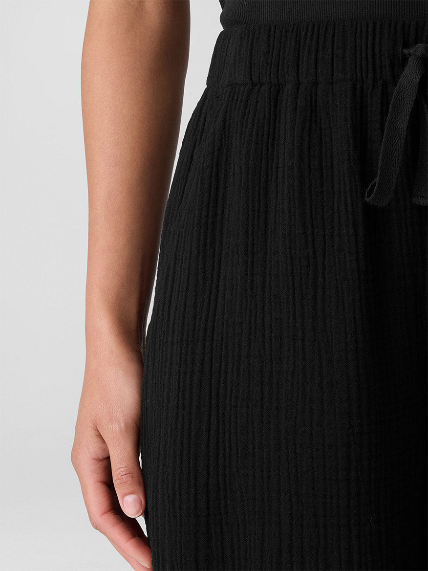 A person wears the Eileen Fisher Organic Cotton Lofty Gauze Wide-Leg Pant in Black, featuring a textured fabric, elastic waistband, and drawstring. The image shows the pant from waist to mid-thigh with only one arm and hand visible.
