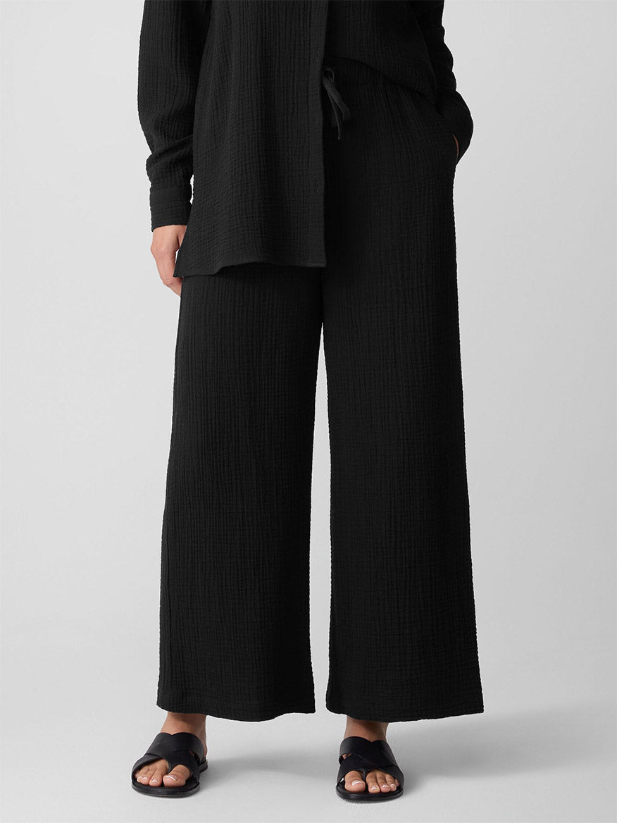 A person stands against a plain background, wearing a loose black textured long-sleeve top and Eileen Fisher Organic Cotton Lofty Gauze Wide-Leg Pant in Black, paired with black sandals.