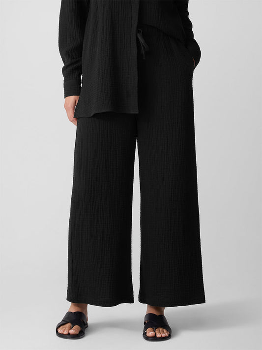 A person stands against a plain background, wearing a loose black textured long-sleeve top and Eileen Fisher Organic Cotton Lofty Gauze Wide-Leg Pant in Black, paired with black sandals.
