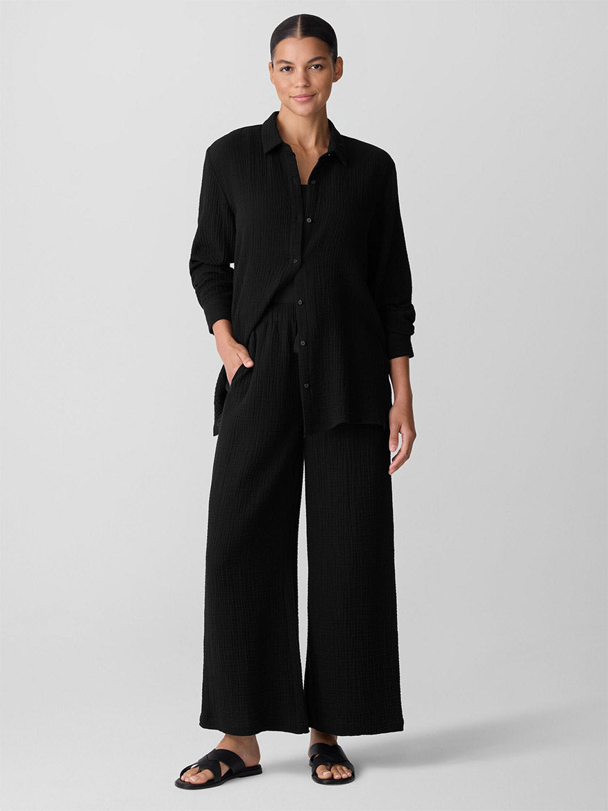 A woman stands against a plain background wearing the Eileen Fisher Organic Cotton Lofty Gauze Wide-Leg Pant in Black with a loose black top and black sandals. Hands in pockets, she smiles slightly.