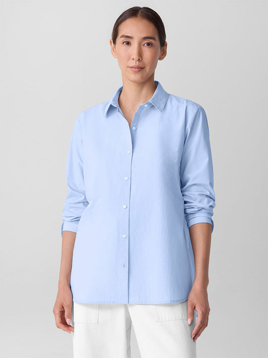 A woman with dark hair in a ponytail wears the Eileen Fisher Striped Poplin Classic Collar Shirt in Myrtle with white pants, standing against a plain light background.