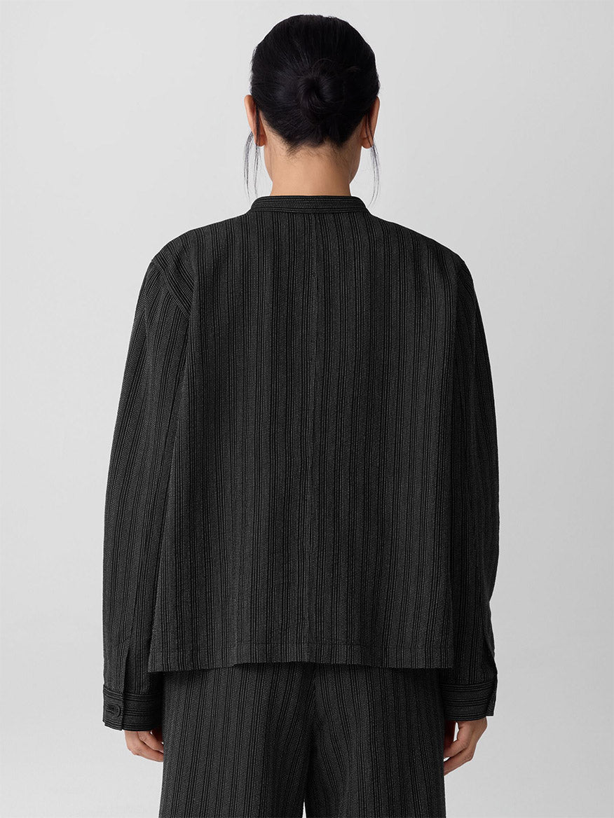 A person with dark hair in a low bun is seen from behind, wearing the Eileen Fisher Cotton Blend Stripe Stitch Shirt Jacket in Black, paired with matching pants against a light gray background.
