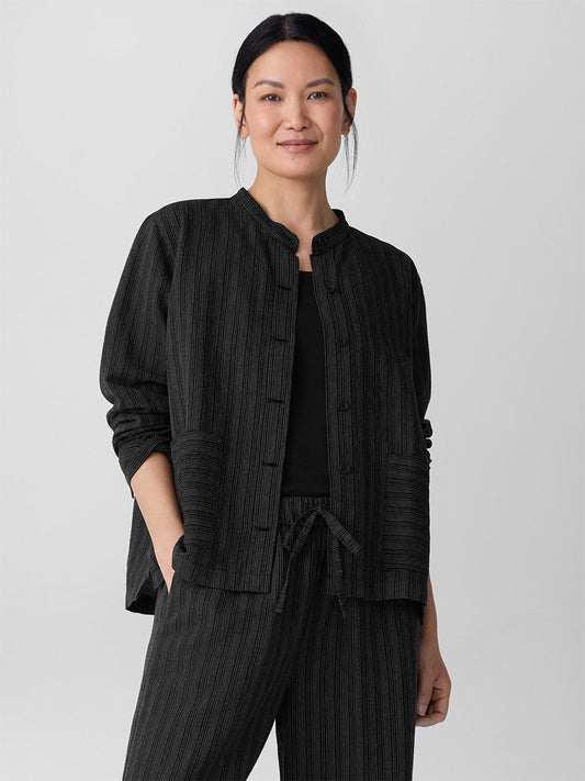 A woman stands against a plain background wearing the Eileen Fisher Cotton Blend Stripe Stitch Shirt Jacket in black, styled with matching pants and her hands in her pockets.