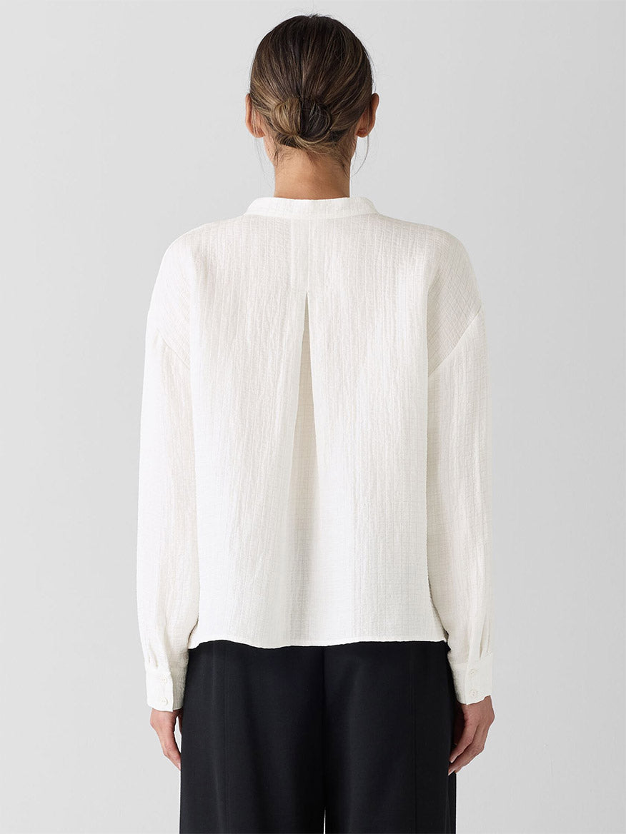 The individual, clad in the Eileen Fisher Mandarin Collar Top in Ivory and black pants, is seen from behind against a plain background. The shirt's mother-of-pearl buttons add subtle elegance, and their hair is tied back neatly.