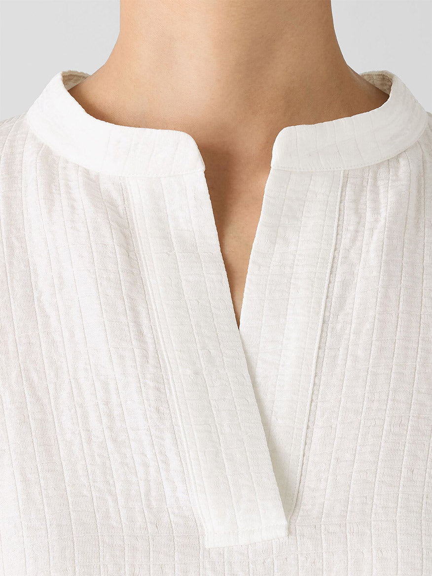 Close-up of someone wearing an Eileen Fisher Mandarin Collar Top in Ivory, a silk and linen shirt with textured fabric, a band collar, and an elegant V-neck, embellished with shimmering mother-of-pearl buttons.