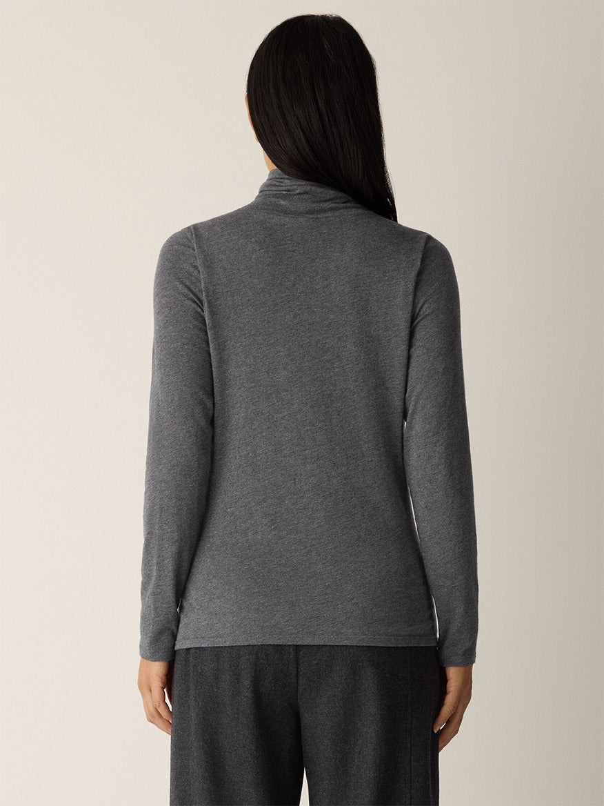 Person shown from behind wears the Eileen Fisher Mélange Sheer Cotton Blend Knit Turtleneck Top in Charcoal, a long-sleeve gray style made of cotton and Tencel Lyocell, ideal for layering over dark pants against a plain light background.