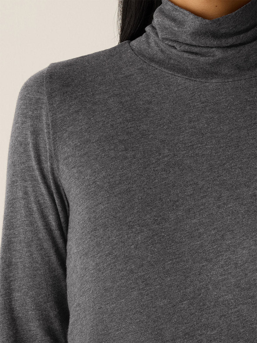 A close-up shows a person wearing the Eileen Fisher Mélange Sheer Cotton Blend Knit Turtleneck Top in Charcoal; only the lower face, neck, and upper torso are visible.