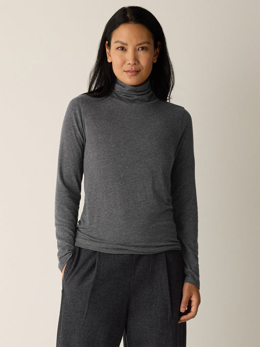 A woman faces forward, wearing the Eileen Fisher Mélange Sheer Cotton Blend Knit Turtleneck Top in Charcoal, paired with dark pants against a plain light background.