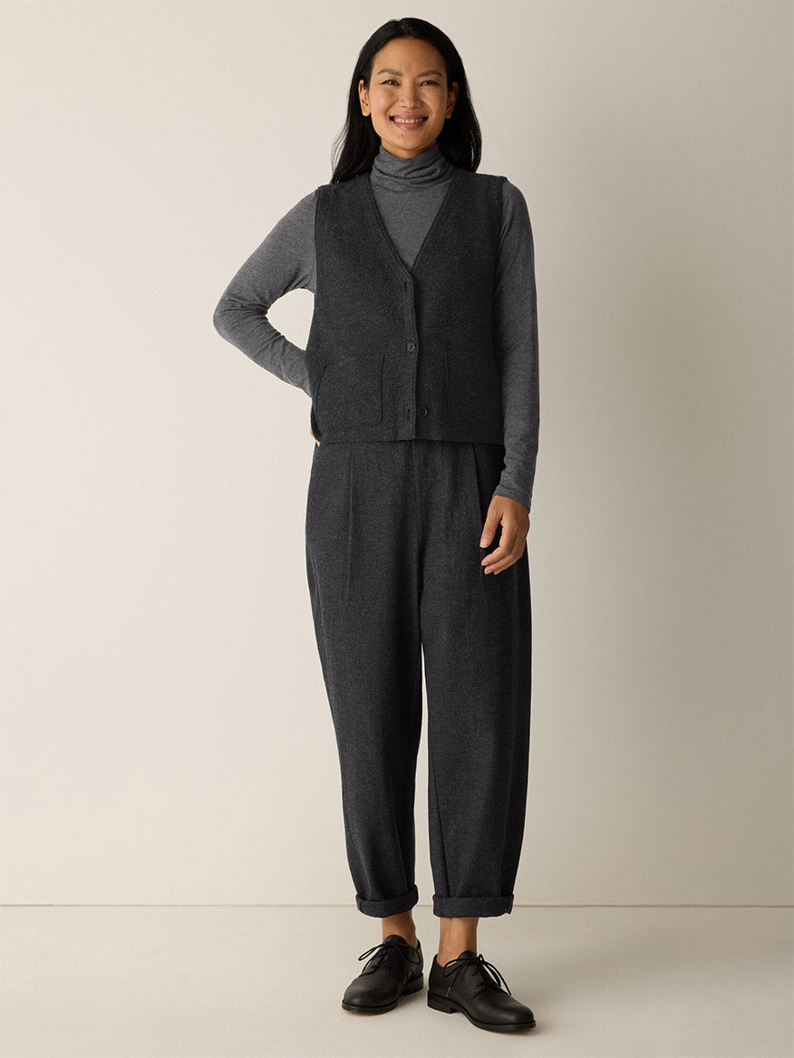 A woman stands indoors against a plain wall, smiling with one hand on her hip. She wears the Eileen Fisher Mélange Sheer Cotton Blend Knit Turtleneck Top in Charcoal, layered under a dark vest with loose dark pants and black shoes.