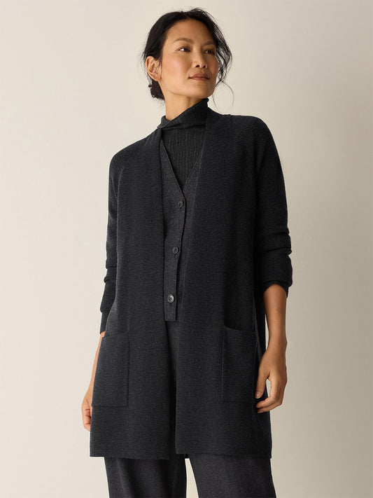 A person stands against a light background wearing the Eileen Fisher Merino Long Cardigan in Charcoal, paired with a matching sweater and pants, with hands in the cardigan pockets.