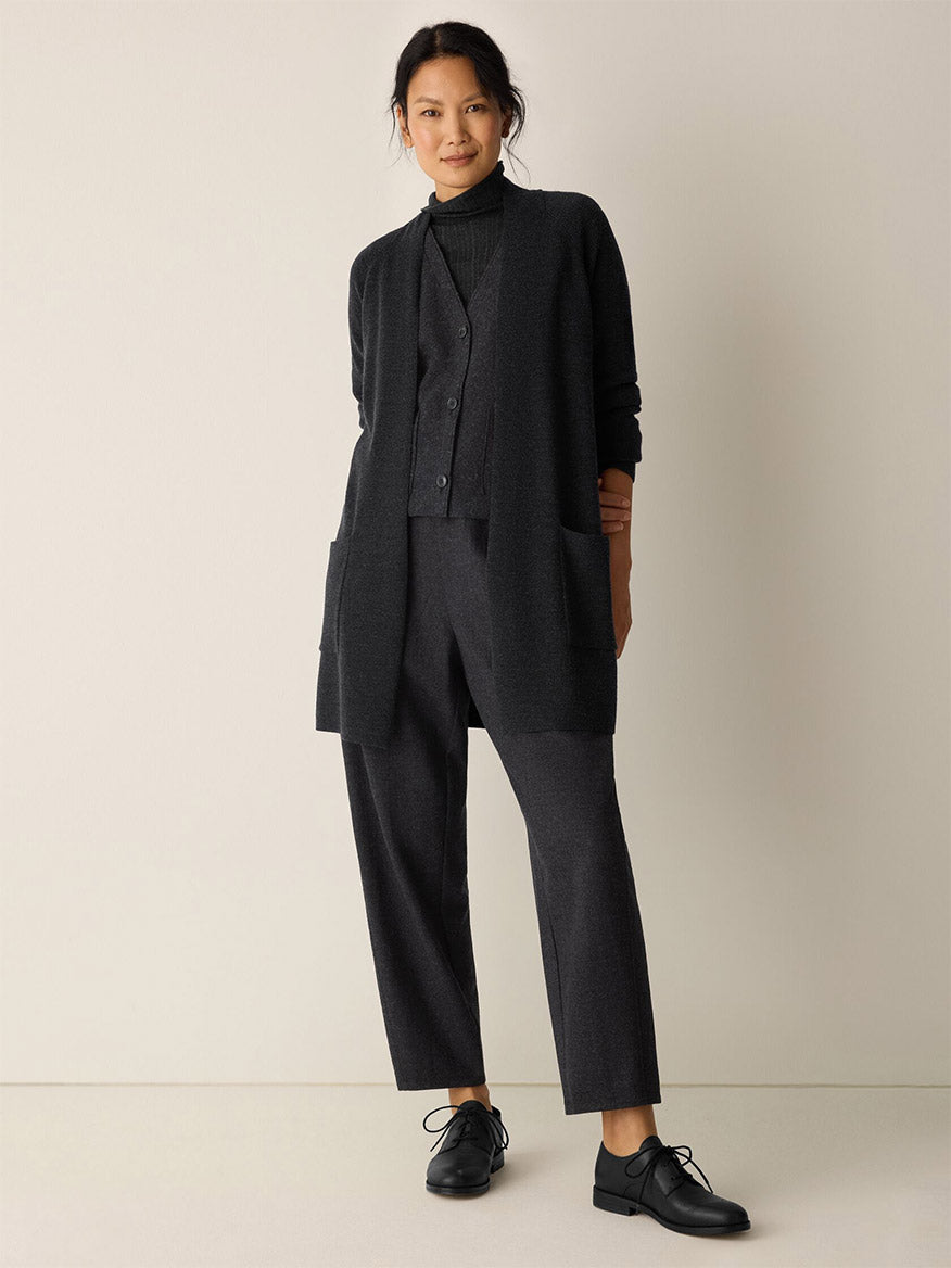 A person stands against a plain background wearing the Eileen Fisher Merino Long Cardigan in Charcoal over coordinated trousers, a buttoned vest, turtleneck, and black dress shoes.