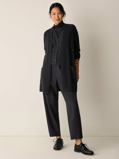 A person stands against a plain background wearing the Eileen Fisher Merino Long Cardigan in Charcoal over coordinated trousers, a buttoned vest, turtleneck, and black dress shoes.