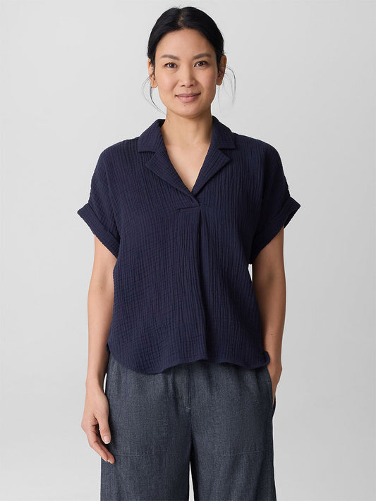 A woman faces the camera, wearing the Eileen Fisher Organic Cotton Lofty Gauze Notch Collar Top in Ink paired with loose-fitting dark gray pants, set against a plain light background.