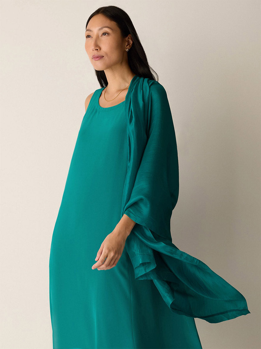 A woman stands against a plain light background, draped in the Eileen Fisher Washed Organic Silk Scarf in Teal over her sleeveless dress.