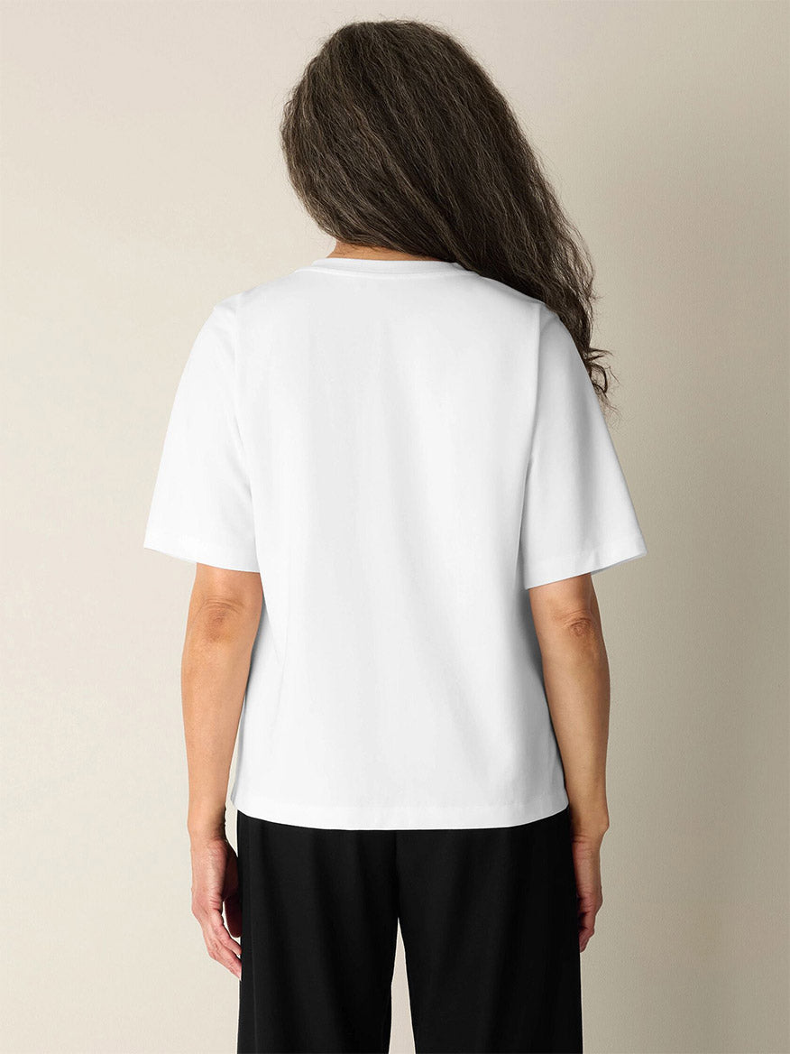 A person with long, wavy brown hair stands facing away, wearing the Eileen Fisher Extra-Fine Ponte Crew Neck Tee in White—crafted from the lightest ponte and Fair Trade Certified materials—and black pants against a neutral background.