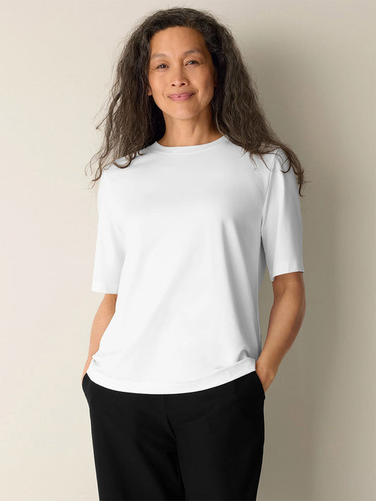 A person with long wavy hair stands against a plain wall, wearing the Eileen Fisher Extra-Fine Ponte Crew Neck Tee in White and black pants, hands in pockets and a neutral expression.
