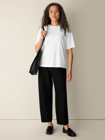 A woman stands against a plain wall, wearing the Eileen Fisher Extra-Fine Ponte Crew Neck Tee in White, paired with black pants, black loafers, and carrying a black shoulder bag.