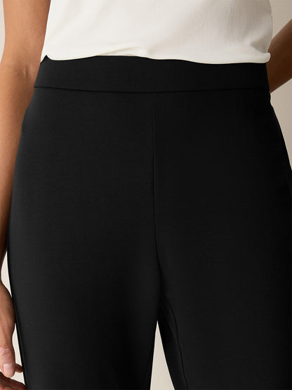 Close-up of a person wearing the Eileen Fisher Extra-Fine Ponte Kick Pant in Black, crafted from our lightest ponte fabric and paired with a white top, highlighting the waistband and upper thigh area with one hand resting by their side. The pants’ subtle flare adds a modern touch to this sleek look.