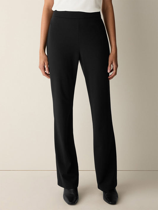 Person standing against a neutral background, wearing the Eileen Fisher Extra-Fine Ponte Kick Pant in Black, crafted from the lightest ponte fabric, paired with black shoes. Their hands are relaxed at their sides. Upper body is partially visible and the top appears white.
