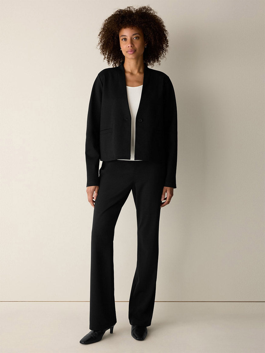 A woman stands against a plain background wearing the Eileen Fisher Extra-Fine Ponte Kick Pant in Black—crafted from the lightest ponte and featuring a subtle flare at the hem—paired with a black blazer, white top, and sleek black shoes.